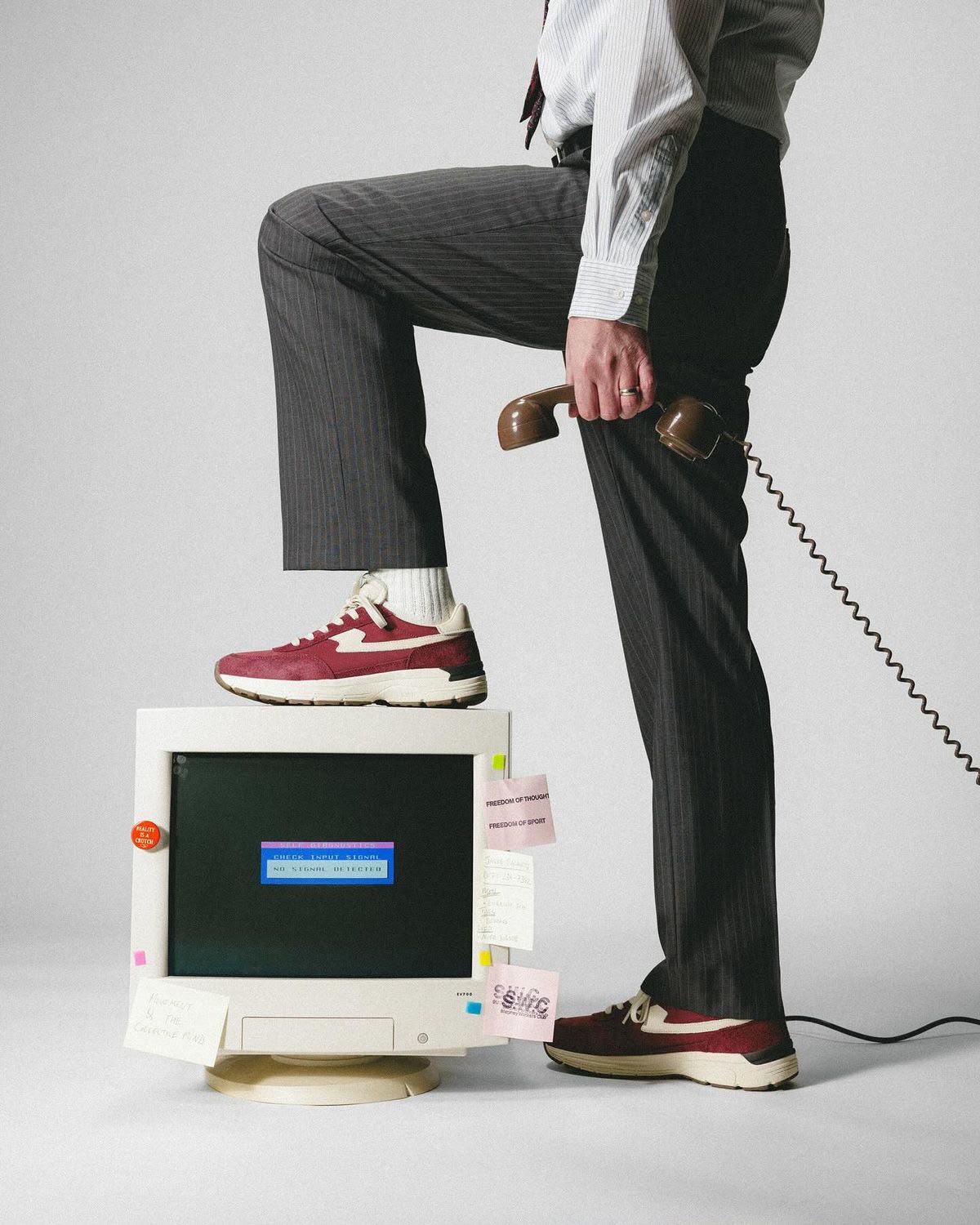 Persona trajeada con un pie apoyado sobre un monitor CRT que muestra NO SIGNAL DETECTED, sosteniendo un telefono retro